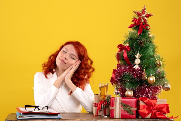 female worker sitting behind table with christmas presents and tree on yellow