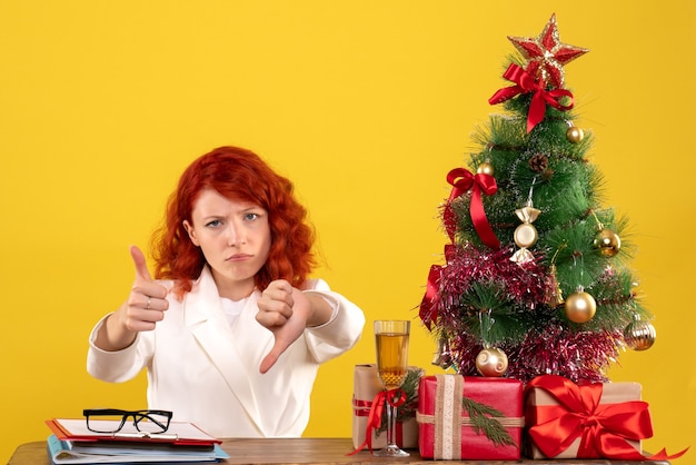 Free photo female worker sitting behind table with christmas presents and tree on yellow