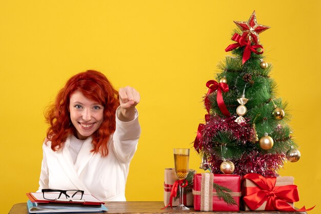 Lavoratrice seduta dietro il tavolo con albero di natale e regali su giallo