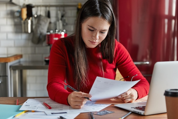 Female worker studies market analysis, makes financial forecast, poses at workplace with documentation, electronic device, works on kitchen.