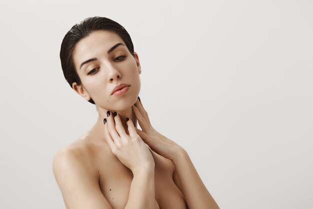 Feminine dreamy woman standing naked in shower, apply skincare routine product