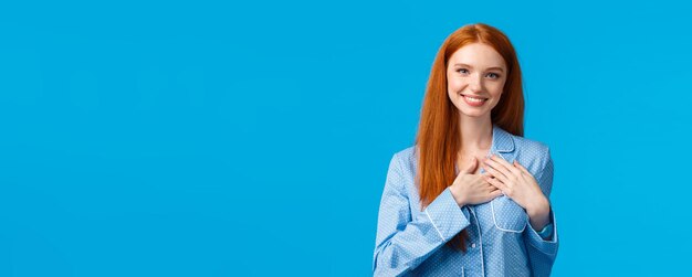 Женственная милая рыжая женщина с длинными рыжими волосами в пижаме держит руки на сердце и улыбается