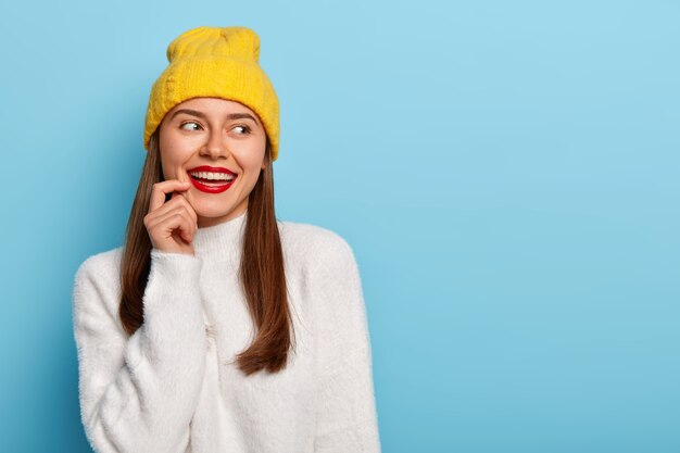 Feminine woman with dark hair, wears red lipstick, wears yellow hat and white soft sweater, smiles broadly, keeps finger near lips, looks aside, stands against blue background.