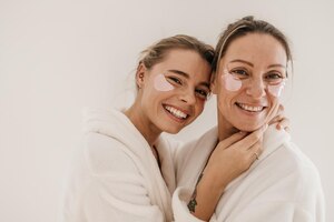 Free photo feminine young caucasian models wear bathrobes after shower and mask for bruising under eyes in spa. skin care and health concept