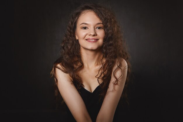 Femininity, beauty and body language. Isolated shot of cute pretty girl with curly dark hair and make up sitting indoors in closed posture