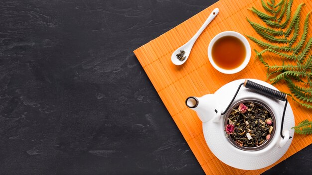 Листья папоротника и сушеные чайные травы с чайником на оранжевой подставке для столовых приборов на черном фоне