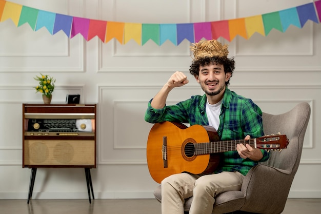 Симпатичный молодой парень Festa junina в соломенной шляпе с ретро-радио и разноцветными флагами, играющий на гитаре