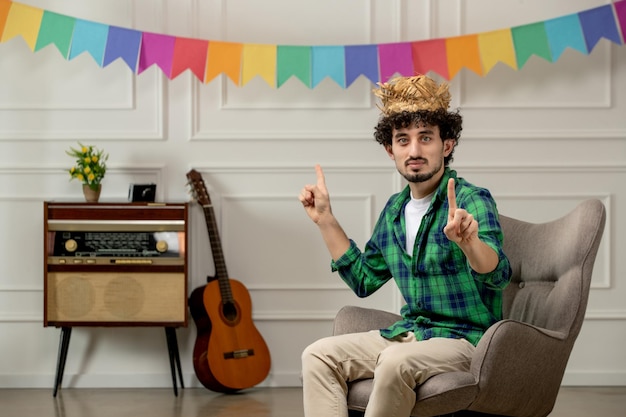 Festa junina cute young guy in straw hat with retro radio and colorful flags pointing fingers up