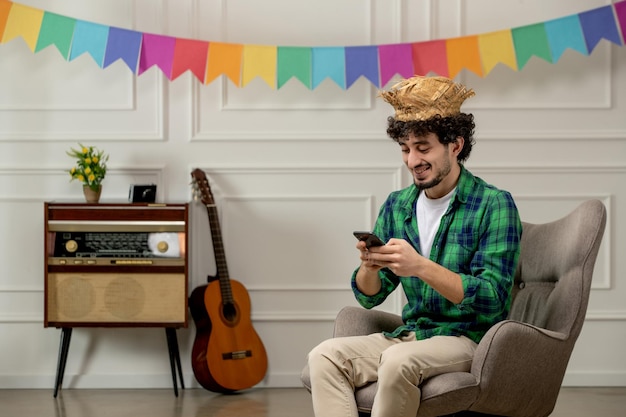 Festa junina cute young guy in straw hat with retro radio and colorful flags typing on the phone