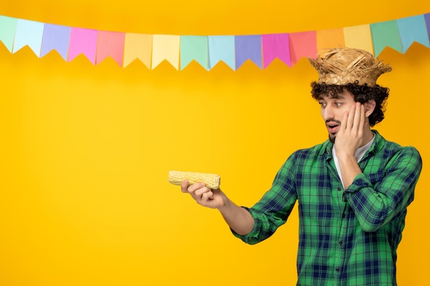 Festa junina молодой симпатичный парень в соломенной шляпе и разноцветных флагах бразильского фестиваля удивил кукурузой