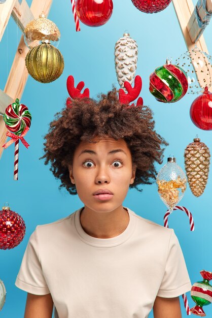 Festive atmosphere in room. Stunned dark skinned woman with curly hair stares bugged eyes hears shocking news wears casual t shirt decorates house for Christmas. Happy holidays time at home.
