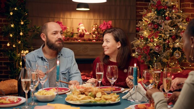 Праздничная пара празднует Рождество за вечерним обеденным столом с близкими членами семьи. Позитивные люди сидят в гостиной, чтобы отпраздновать настоящий традиционный зимний праздник с близкими.