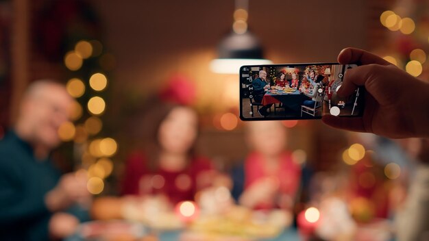 Festive family members getting photographed with smartphone while sitting at table enjoying Christmas dinner together. Person taking photo of happy people celebrating winter holiday with phone device.