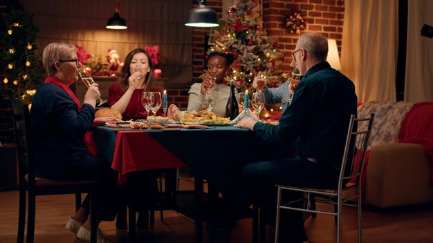 Festive happy woman toasting with family members while enjoying home cooked Christmas dinner at home. Joyful multiethnic people celebrating traditional winter holiday while drinking sparkling wine.