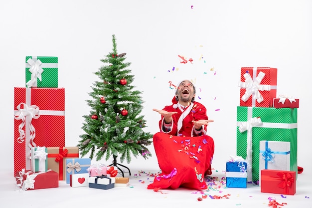 Festive holiday mood with happy santa claus sitting on the ground and playing with christmas decorations near gifts and decorated xsmas tree on white background