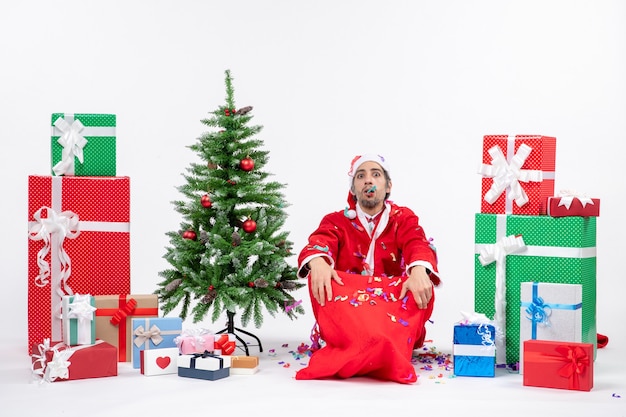 Festive holiday mood with sad santa claus sitting on the ground and playing with christmas decorations near gifts and decorated xsmas tree on white background