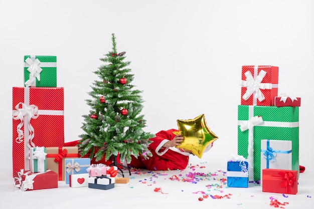 Festive holiday mood with santa claus lying behind christmas tree closing his face near gifts in different colors on white background