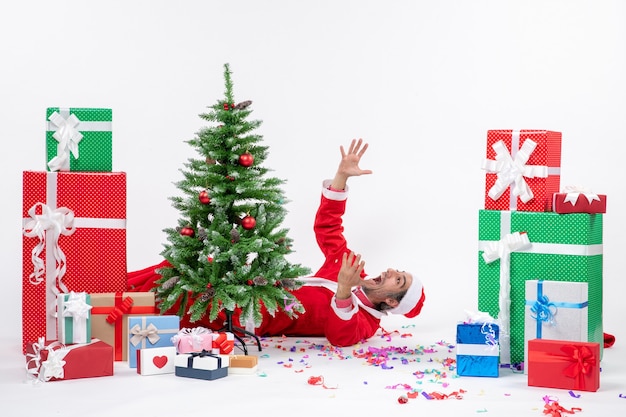 Festive holiday mood with young nervous santa claus lying behind christmas tree near gifts on white background