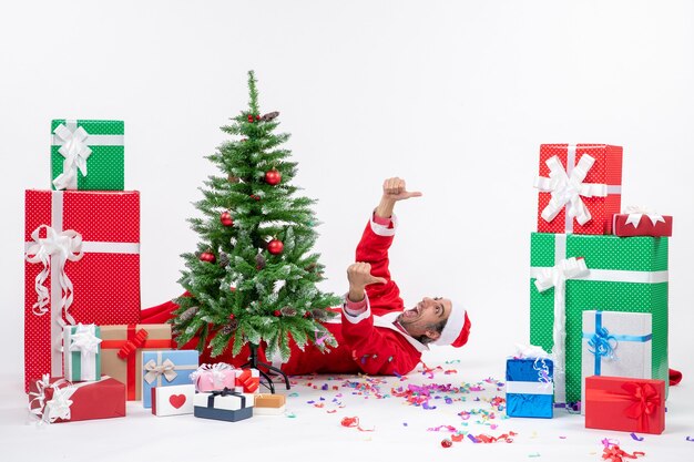 白い背景の上の贈り物の近くのクリスマスツリーの後ろに横たわっている若いポジティブなサンタクロースとお祝いの休日の気分