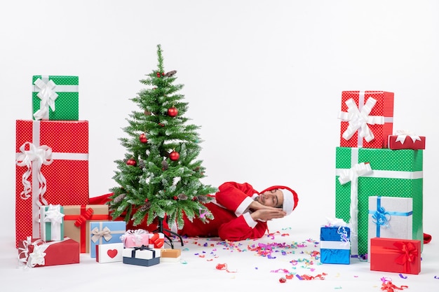 Foto gratuita umore di festa festiva con il giovane babbo natale sdraiato dietro l'albero di natale vicino a doni in diversi colori su sfondo bianco stock photo