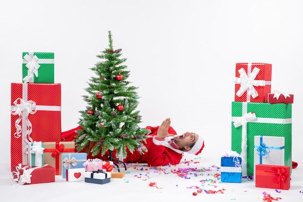 Festive holiday mood with young shocked santa claus lying behind christmas tree near gifts on white background