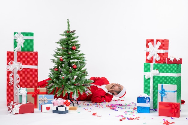 白い背景の上の贈り物の近くのクリスマスツリーの後ろに横たわっている若い驚きのサンタクロースとお祝いの休日の気分