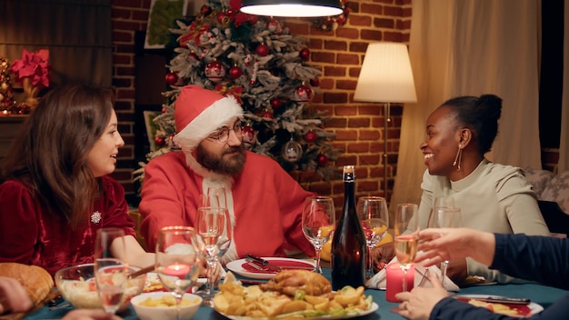 크리스마스 저녁 파티에서 산타클로스 의상을 입고 가족과 이야기하는 축제 남자. 겨울 휴가를 축하하면서 사람들과 토론하는 산타클로스로 변장한 젊은 성인.