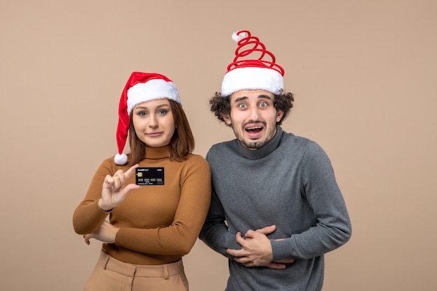 Festive mood with cool couple wearing red santa claus hats girl showing bank card on gray