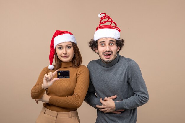 Festive mood with cool couple wearing red santa claus hats girl showing bank card on gray