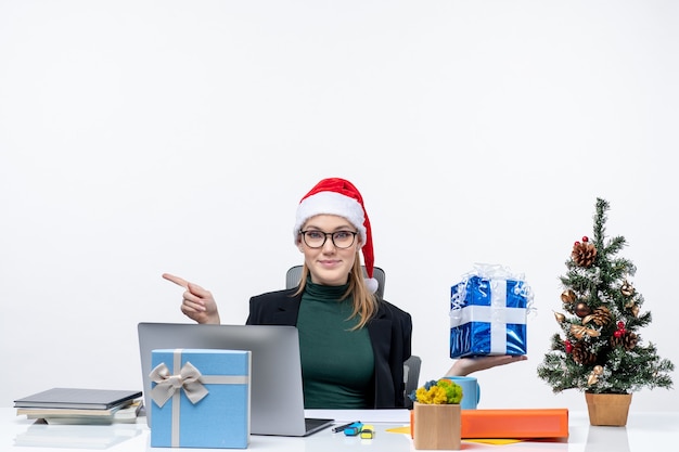 右側に何かを指しているクリスマスプレゼントを示すテーブルに座って眼鏡をかけているサンタクロースの帽子をかぶった好奇心旺盛な驚きの笑顔の若い女性とお祭り気分