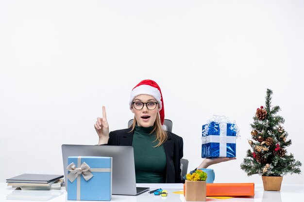 Праздничное настроение с удивленной молодой женщиной в шляпе Санта-Клауса и в очках, сидящей за столом, показывающим рождественский подарок, указывающим выше на белом фоне