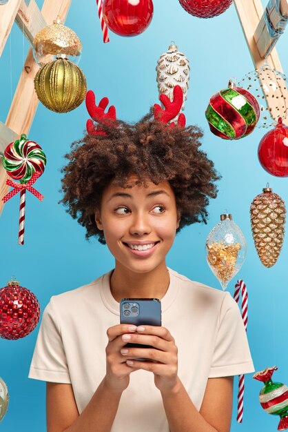 Festive spirit in air. Merry Christmas concept. Glad dark skinned woman uses smartphone to send congratulations for relatives poses in decorated room looks aside smiles happily. New Year is soon