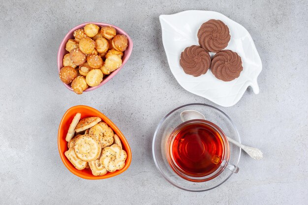 Few cookies on plate next to bowls of cookie chips and a cup of tea on marble surface.