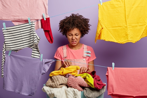 Fie, what dirty laundry! Upset Afro American female looks with dissatisfaction at pile of clothes for washing, frowns face from stench, poses near clotheslines, fed up of housework and cleaning