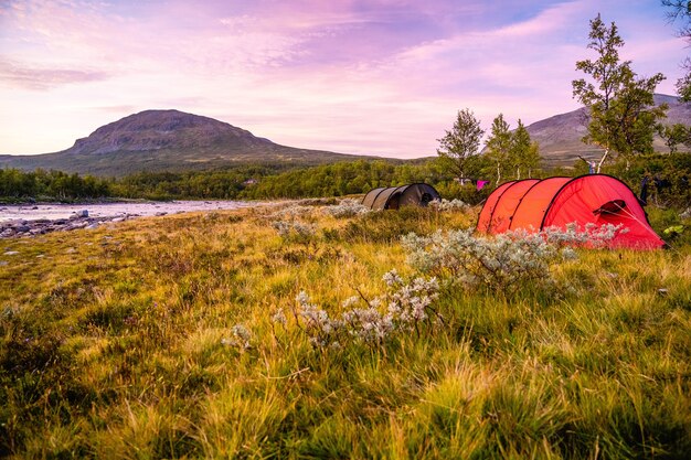 Field with tents surrounded by hills covered in greenery under a cloudy sky during the sunset