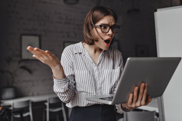 Fierce business lady looks indignantly and shocked into laptop and spreads her arms against background of white workplace.