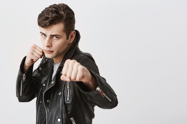 Fierce and confident european male model in black leather jacket with trendy haircut holding fists in front as if ready for fight or challenge, pursuing lips, with determined expression on face