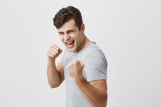 Fierce and confident male with trendy haircut holding fists in front of him as if ready for fight or any challenge, demonstrating his white teeth in anger, having aggressive expression on his face