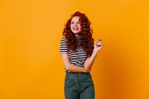 Fiery redhead girl in moms jeans and striped top is smiling and posing on yellow space.