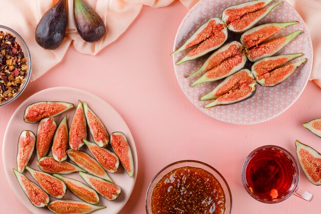 Figs in plates with cup of tea, jam, dried herbs flat lay on pink and textile