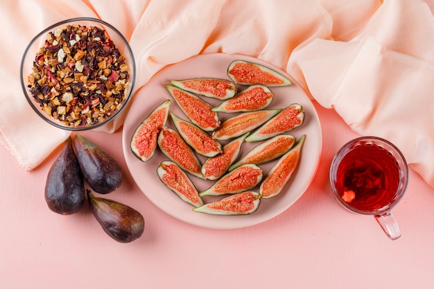 Free photo figs with cup of tea, dried herbs in a plate on pink and textile, flat lay.