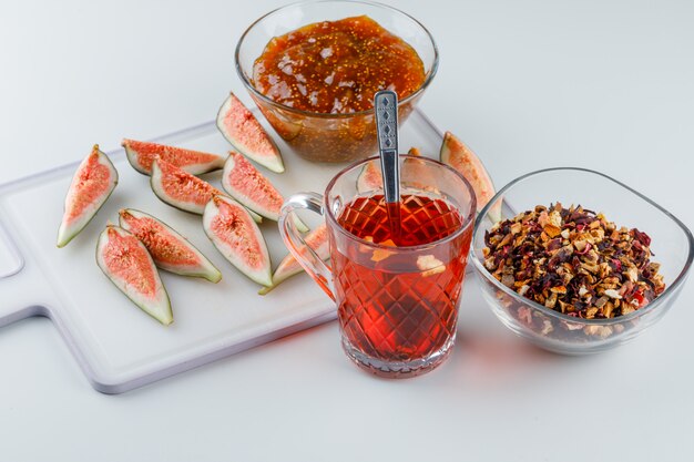 Figs with fig jam, tea, teaspoon, dried herbs on white and cutting board, high angle view.