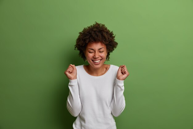 Finally I gained it! Cheerful successful woman raises hands with excitement, clenches fists in triumph, cheers with beaming smile and closed eyes, rejoices winning prize, stands indoor over green wall