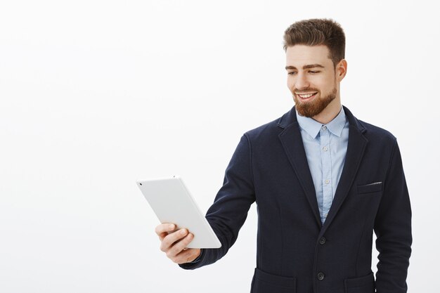 Finance, gig economy and business concept. Self-assured delighted successful male entrepreneur in elegant stylish suit holding digital tablet gazing at gadget screen satisfied with assured smile