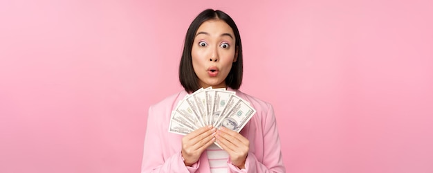 Финансовый микрокредит и концепция людей Счастливая улыбающаяся азиатская деловая женщина, показывающая доллары, стоящие в костюме на розовом фоне