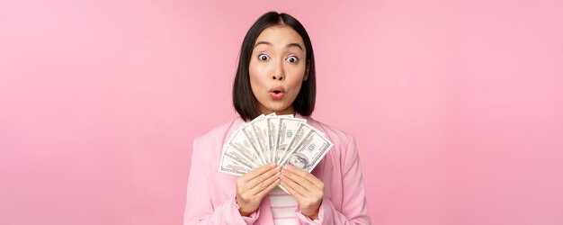金融マイクロクレジットと人々の概念ピンクの背景にスーツで立っているドルのお金を示す幸せな笑顔のアジアの実業家