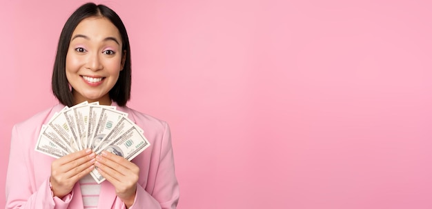 Finance microcredit and people concept Happy smiling asian businesswoman showing dollars money standing in suit against pink background