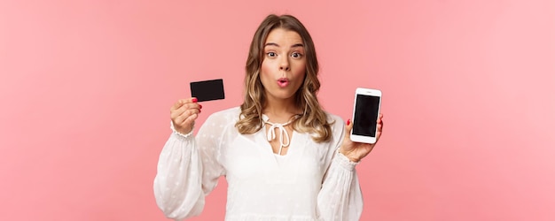 금융 쇼핑 및 기술 개념 하얀 드레스를 입은 흥분한 금발의 귀여운 소녀의 클로즈업 초상화는 신용 카드와 휴대 전화 분홍색 배경을 보여주는 것처럼 카메라를 즐겁게 봅니다.