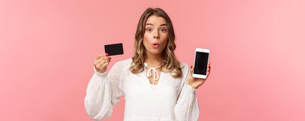 金融ショッピングとテクノロジーのコンセプト白いドレスを着た興奮した金髪のかわいい女の子のクローズアップの肖像画は、クレジットカードと携帯電話のピンクの背景を示すようにカメラを面白がって折ります