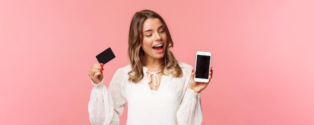 Finance shopping and technology concept Closeup portrait of excited goodlooking blond girl in white dress renew her spring wardrobe with buying clothes online via smartphone hold credit card